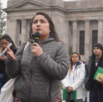 Dulce Gutierrez with a microphone, standing in front of a line of people with signs in front of a building