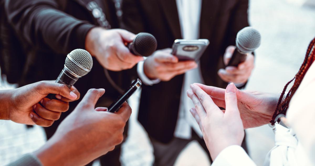 A group of reporters hold up microphones and cellphones, reporters and interviewees faces are obscured.