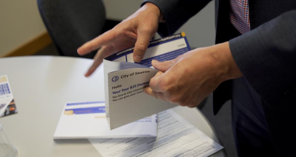 A man's hands opening an envelope that says "City of Seattle. Hello, your four $25 Democracy Vouchers..."