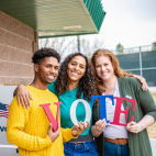 A picture of three people holding letters that spell VOTE. They are standing close together in front of a polling location.