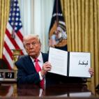Donald Trump sitting at a desk in the Oval Office holding up an executive order with his signature on it.