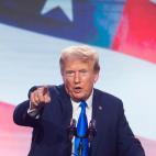 Donald Trump speaking while pointing at the camera in front of a giant American flag
