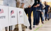 A table with signs that say "Vote" and a line of people out of focus in the background