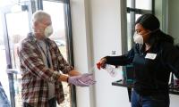 A man wearing a medical mask holds out gloved hands while a woman wearing a medical mask sprays his hands.