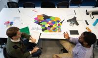 A bird's eye view of a map of Wisconsin on a table with district lines drawn and two people sitting next to it discussing.