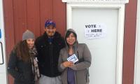 Three people outside a building in the snow. One of the women holds a sign that says "I Voted". A sign on the door to the building says "Vote Here".