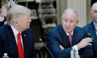 Stephen Schwarzman speaks into a microphone while sitting at a table while President Trump looks on