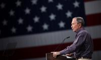 Michael Bloomberg speaking at a podium with an American flag in the background