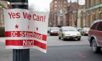 A sign saying "Yes we can! DC Statehood Now!" hanging on a lamppost with traffic going by in the background.
