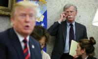 John Bolton leaning against a wall looking on as Trump sits out of focus in the foreground speaking