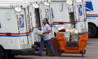 A woman wearing a mask carries a crate to the back of a postal truck