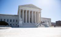 Wide shot of the Supreme Court with blue sky and the sun behind it
