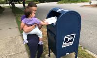 Ruth Greenwood placing her ballot in a blue USPS mailbox while holding her daughter.