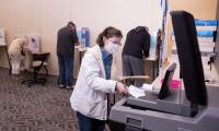 A woman places a ballot into a machine while people behind her fill out ballots in voting booths