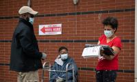 A woman write on a clipboard while another woman looks on from a seated position with a walker in front of her. A man looks on as well. All are wearing face masks.