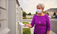 A woman wearing a face mask puts an envelope into a mailbox