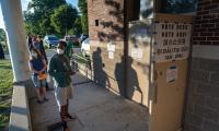 Voters line up along a concrete ramp outside a propped open door with a "Vote Here" sign on it.