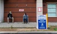 A man and elderly woman wearing masks sit on chairs on a sidewalk with a "Vote Here" sign on the building behind them.