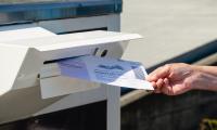 A hand holding a ballot inserts it into a slot in a drop box.
