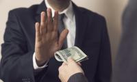 A man in a suit holding up his hand to refuse a wad of money being handed to him by another man off-camera