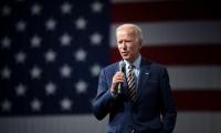 Joe Biden speaking while holding a microphone in front of a large American flag