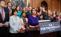 Nancy Pelosi standing a a podium on which is written the words "H.R. 1 For the People Act" with a large group of people standing around and behind her.