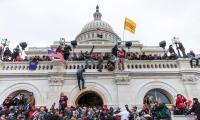 Protestors in a large crowd at the base of the Capitol building as well as climbing up and on top of the wall surrounding it.