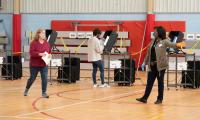 A woman wearing a mask and carrying a ballot walks across the floor of a gym while being directed by another woman who is an election official.
