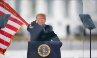 Donald Trump points towards the camera while speaking at a podium behind glass and next to an American flag