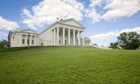 A white state capitol building on top of a green grassy hill under a blue sky with clouds.