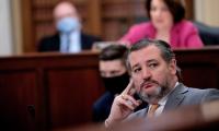 Ted Cruz resting his head on his fingers while sitting at a desk with several people out of focus in the background behind him.