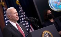 Joe Biden speaking at a podium with an American flag in the background. Kamala Harris out of focus next to him wearing a mask.