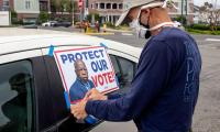 A man wearing a mask tapes a sign to the window of a car. The sign reads "Protect our vote!" and has a picture of John Lewis.