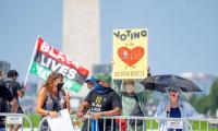 People with sign stand behind a barricade in front of the Washington Monument. One sign reads "Voting is the heart of our democracy". A man in the background carries a Black Lives Matter Flag.