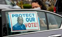 A woman tapes a sign with an image of John Lewis to a car window. The sign reads "Protect Our Vote".