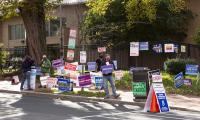 A person holds a sign while sanding among other candidate signs on a sidewalk corner
