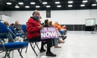 A woman seated in the front row of a group of folding chairs holds a sign which reads "Fair maps now".