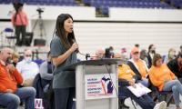 A woman speaking at a podium in a large gymnasium with many audience members behind her.