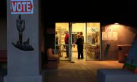 People stand in line, illuminated by light inside the open door of a building. A sign on the side of the building says "Vote" with an image of a cactus beneath it.