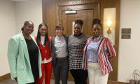 Five women standing in a row in front of a door that says "House Hearing Room".