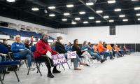 A woman seated in the front row of a group of folding chairs holds a sign which reads "Fair maps now". Others holding signs are seating next to her.