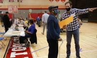 A man gestures with his arm to another man who is holding a large envelope. Behind them is a gymnasium with a row of tables.