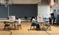 Two women seated at tables facing away from each other in a school gym with cardboard voting booths set up on the tables