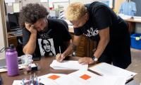 A man and woman lean over a piece of paper on a table while the woman writes on it