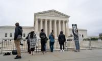 People stand a long a fence in front of the Supreme Court. One woman wearing a poncho holds up a sign which says "It's about US"