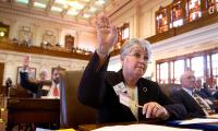 A woman sits at a desk with her hand raised. Others in the room behind her also sit at desks with their hands raised