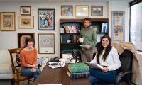 Three people sitting in an office around a table with a bookshelf and photos on the wall behind them