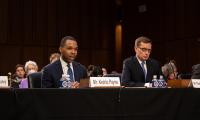 Kedric Payne speaking into a microphone and seated at a table next to another man with audience members behind them.