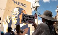 A woman holds a sign which says "Voting Access for All" while standing in front of a large mural of John Lewis.