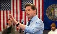 Dean Phillips standing with a microphone in front of an American flag with his hand raised in front of him